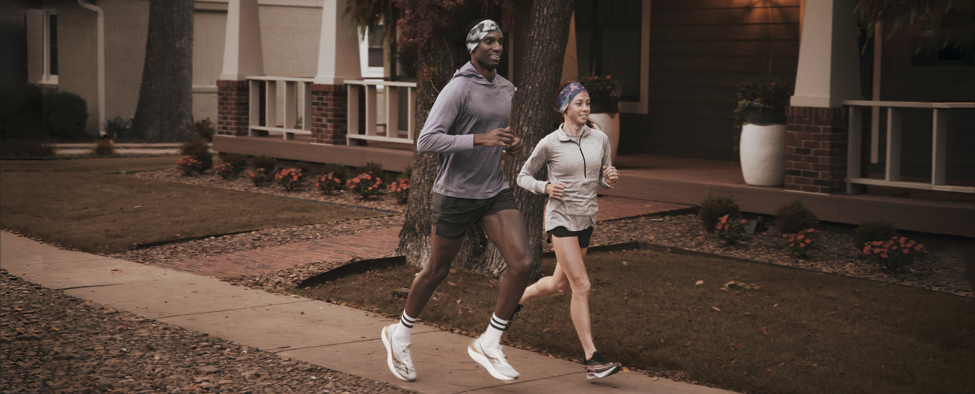 A man and a woman each wearing a JUNK Ear Warmer headband jogging along a residential sidewalk in Fall.